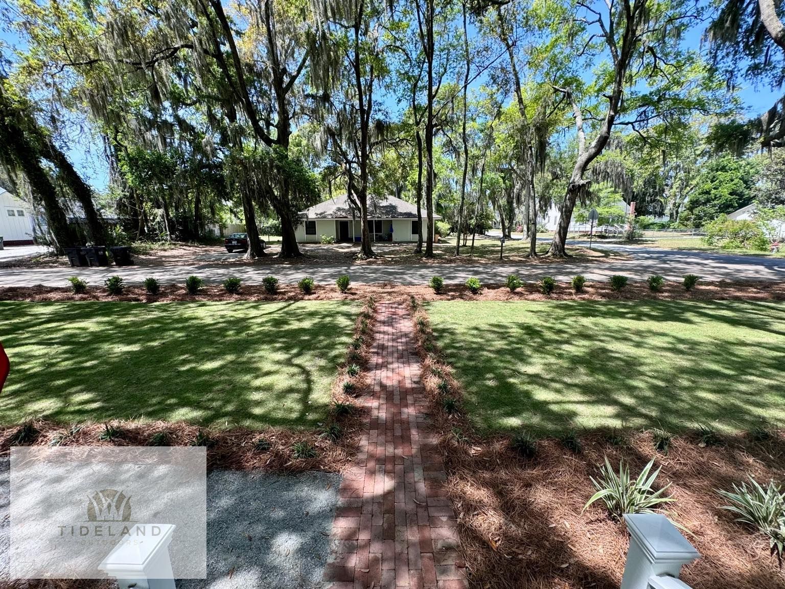 A grassy front yard with a central brick path leading to a distant house surrounded by tall, moss-draped oak trees.
