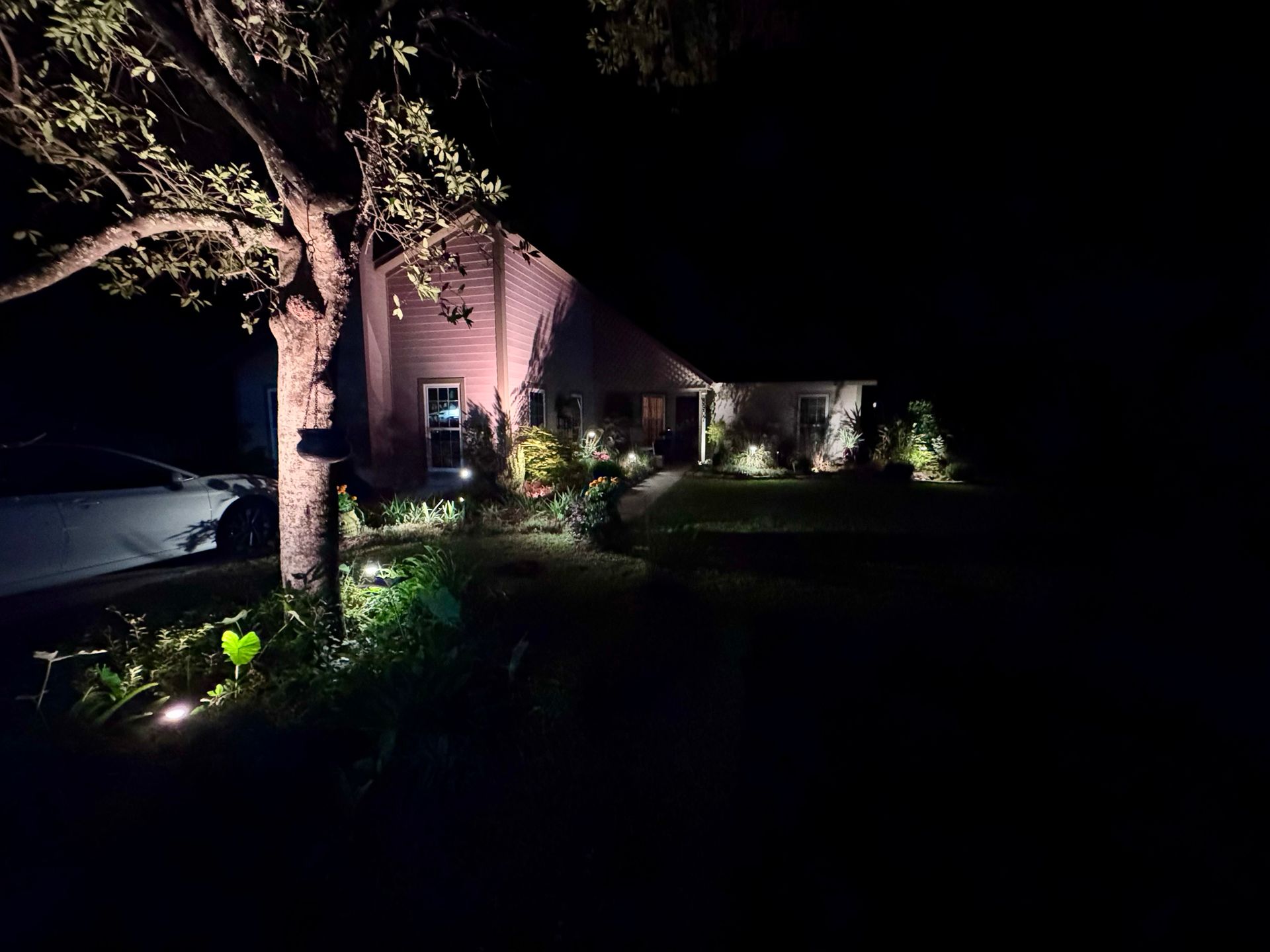 A house exterior at night illuminated by warm landscape spotlights, with a large tree in the foreground.