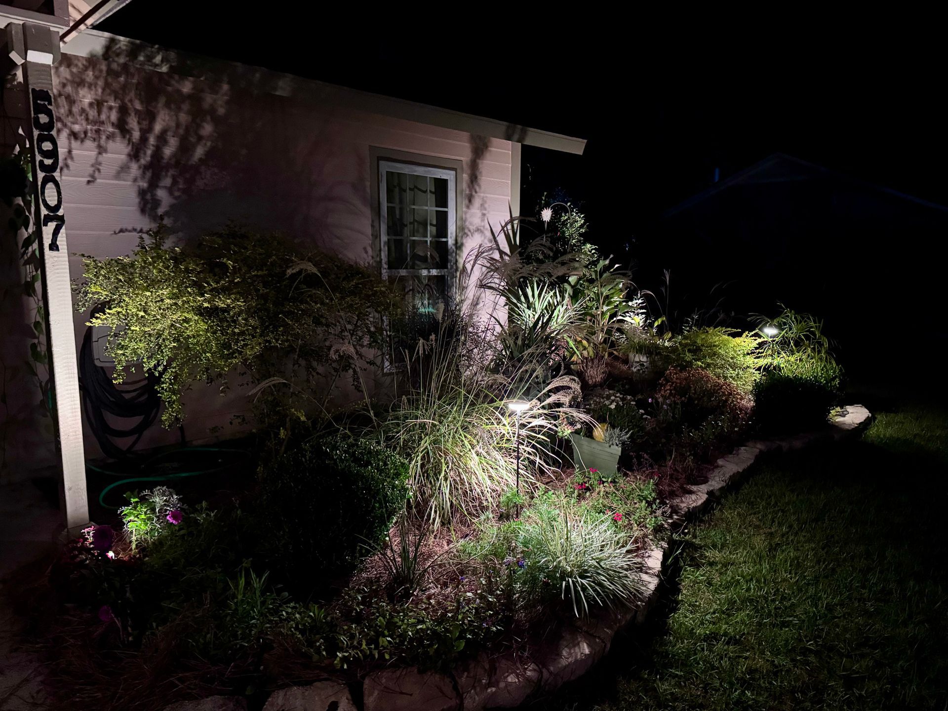 Illuminated house exterior at night with landscaped gardens, stone border, and address number 6907 on a post.