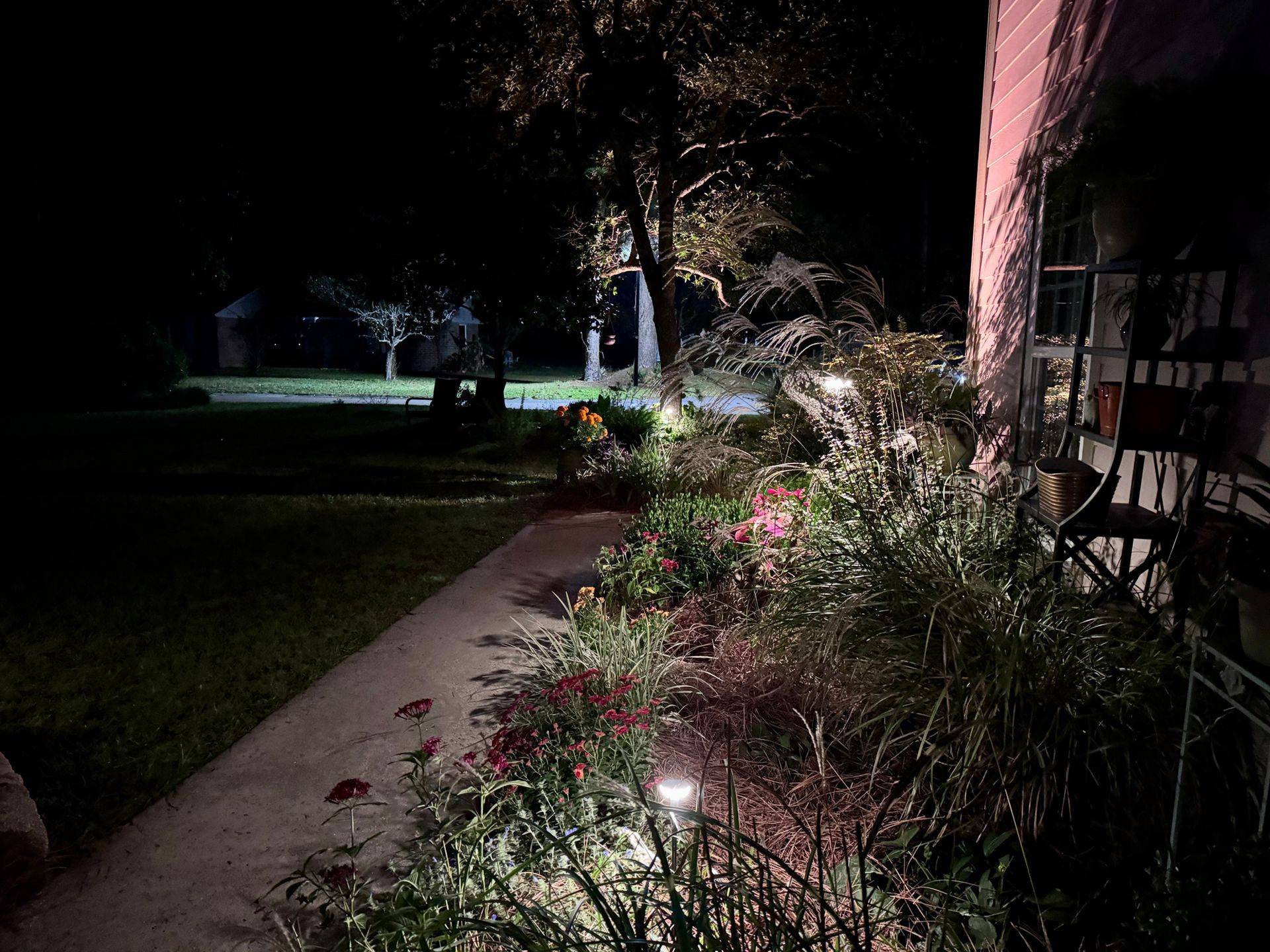 A garden path and landscaping illuminated by landscape lighting at night, with a house structure visible on the right.