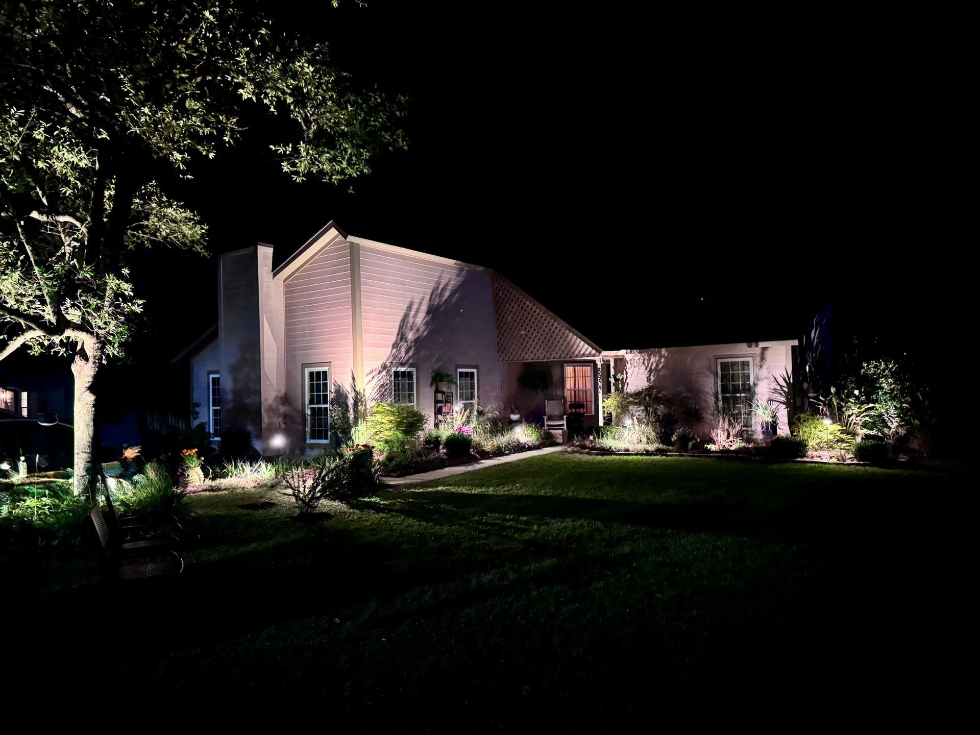 A single-story house at night illuminated by warm landscape lighting, with a large tree in the foreground.