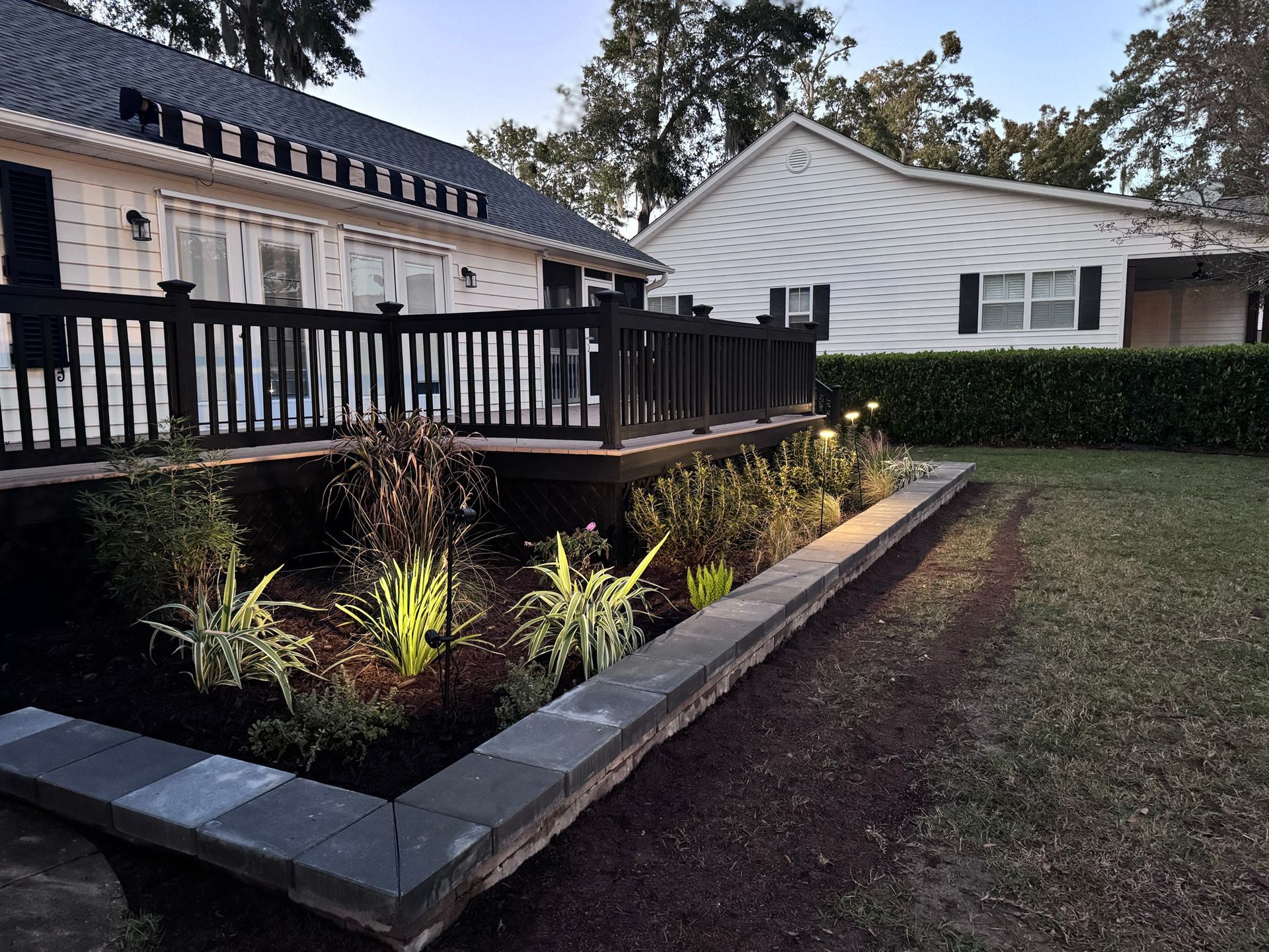A backyard deck with a dark railing overlooks a landscaped garden bed with dark mulch and low-voltage lighting.