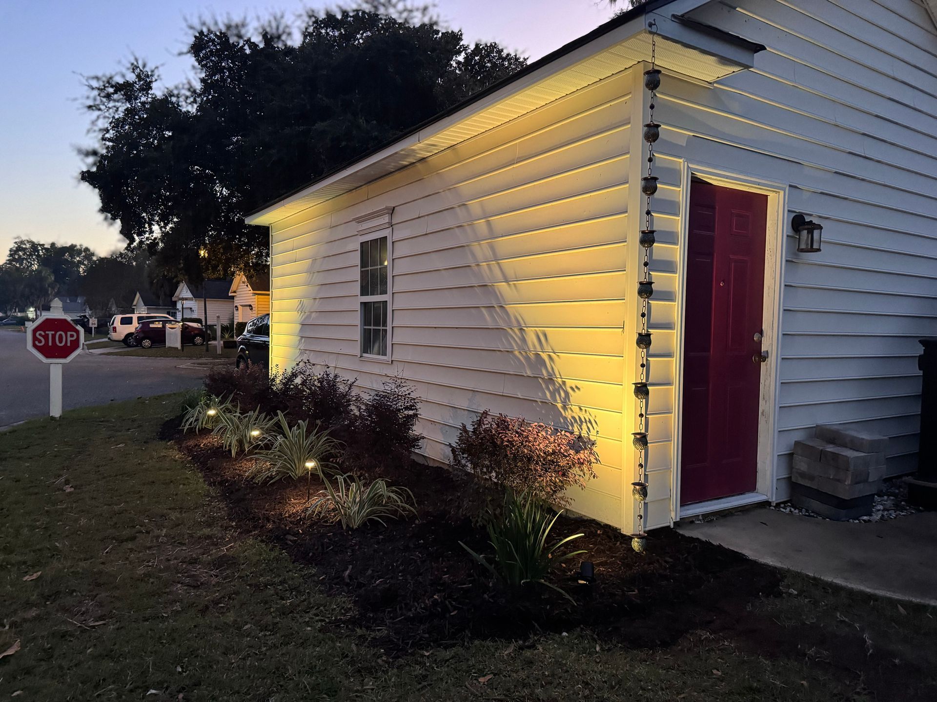 A small white building with a red door at dusk, illuminated by warm exterior lights, with landscaping in the foreground.
