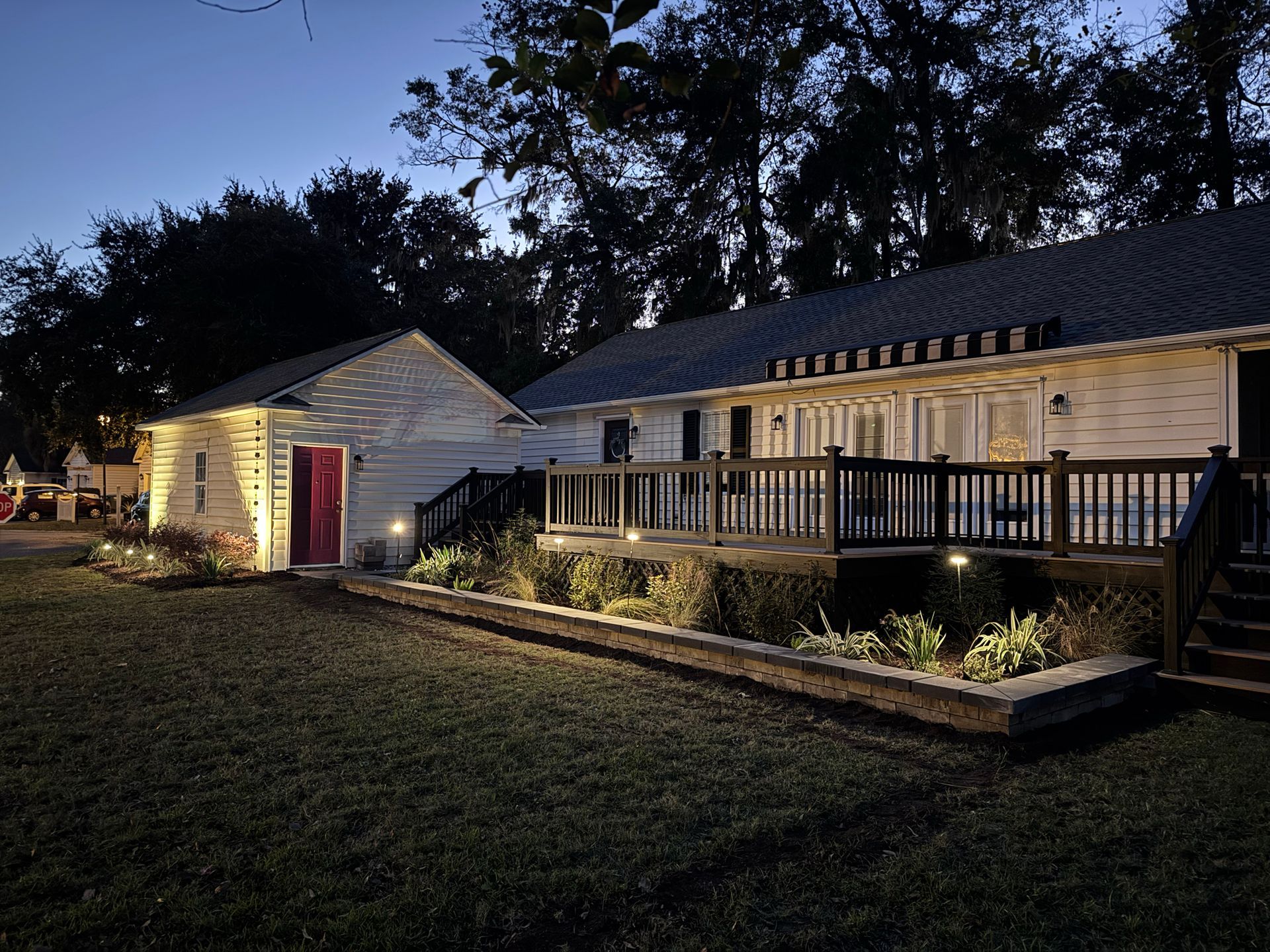 A white house with a wooden deck and a detached garage, illuminated by soft landscape lighting at twilight.