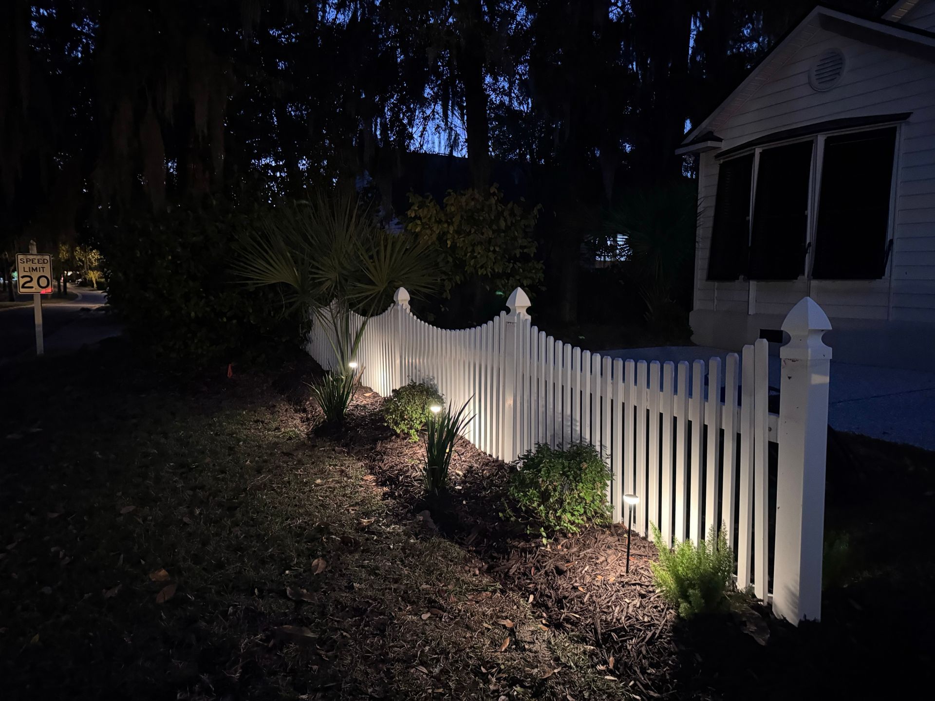 A white picket fence at dusk, illuminated by small solar path lights in a mulched garden bed next to a house.
