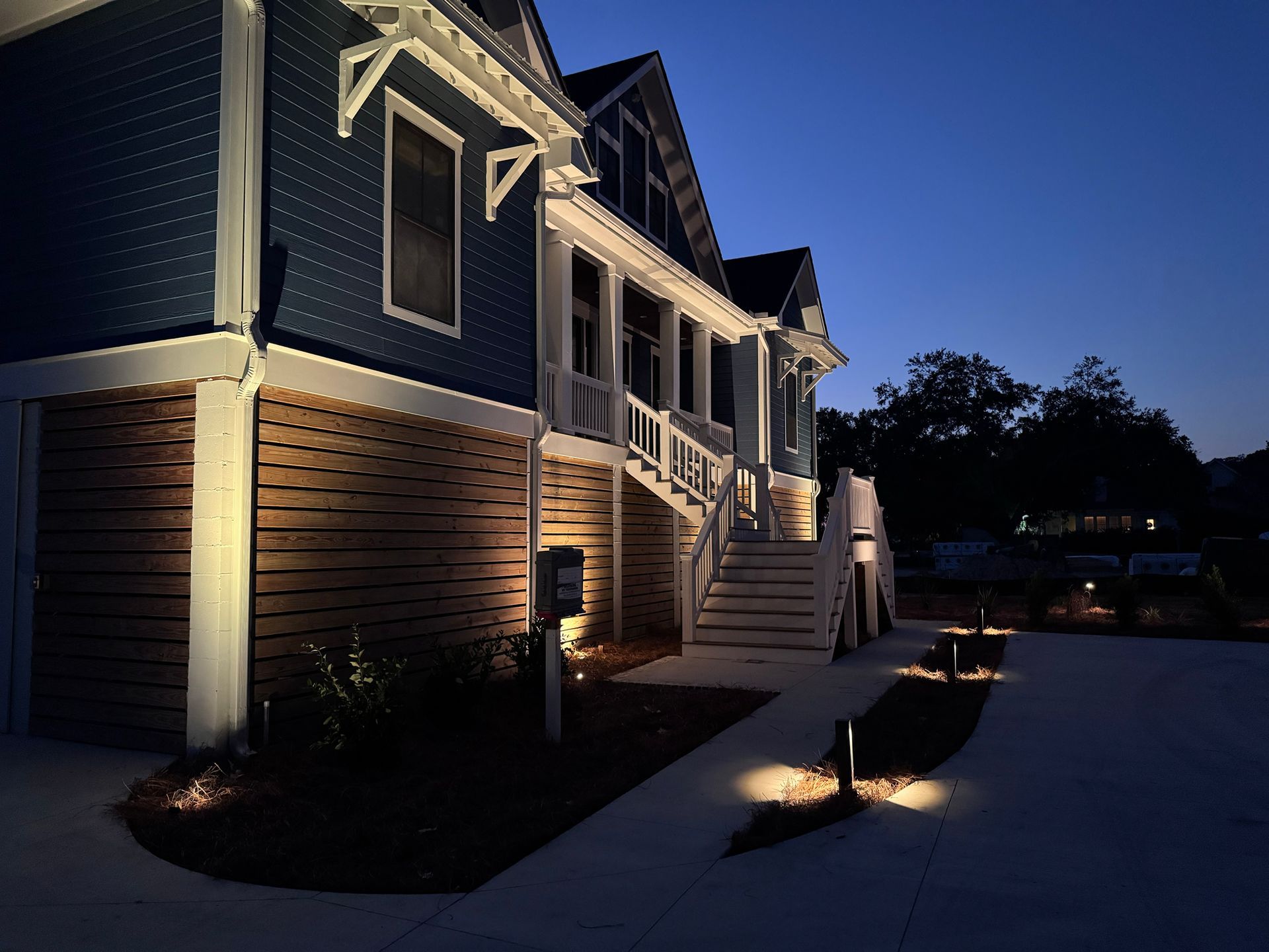 A blue-sided home on stilts illuminated by landscape lighting at twilight, featuring wooden lattice and a staircase.