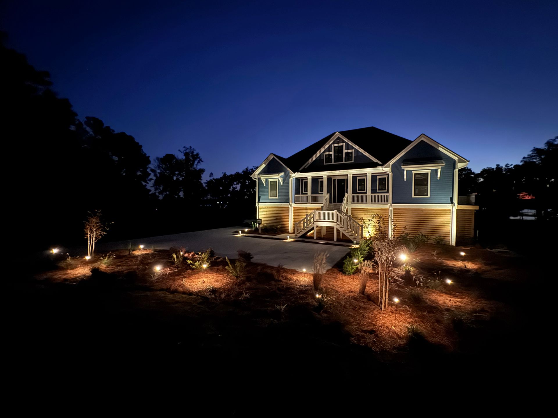 A blue elevated house at night, illuminated by warm landscape lighting in the foreground against a dark blue sky.