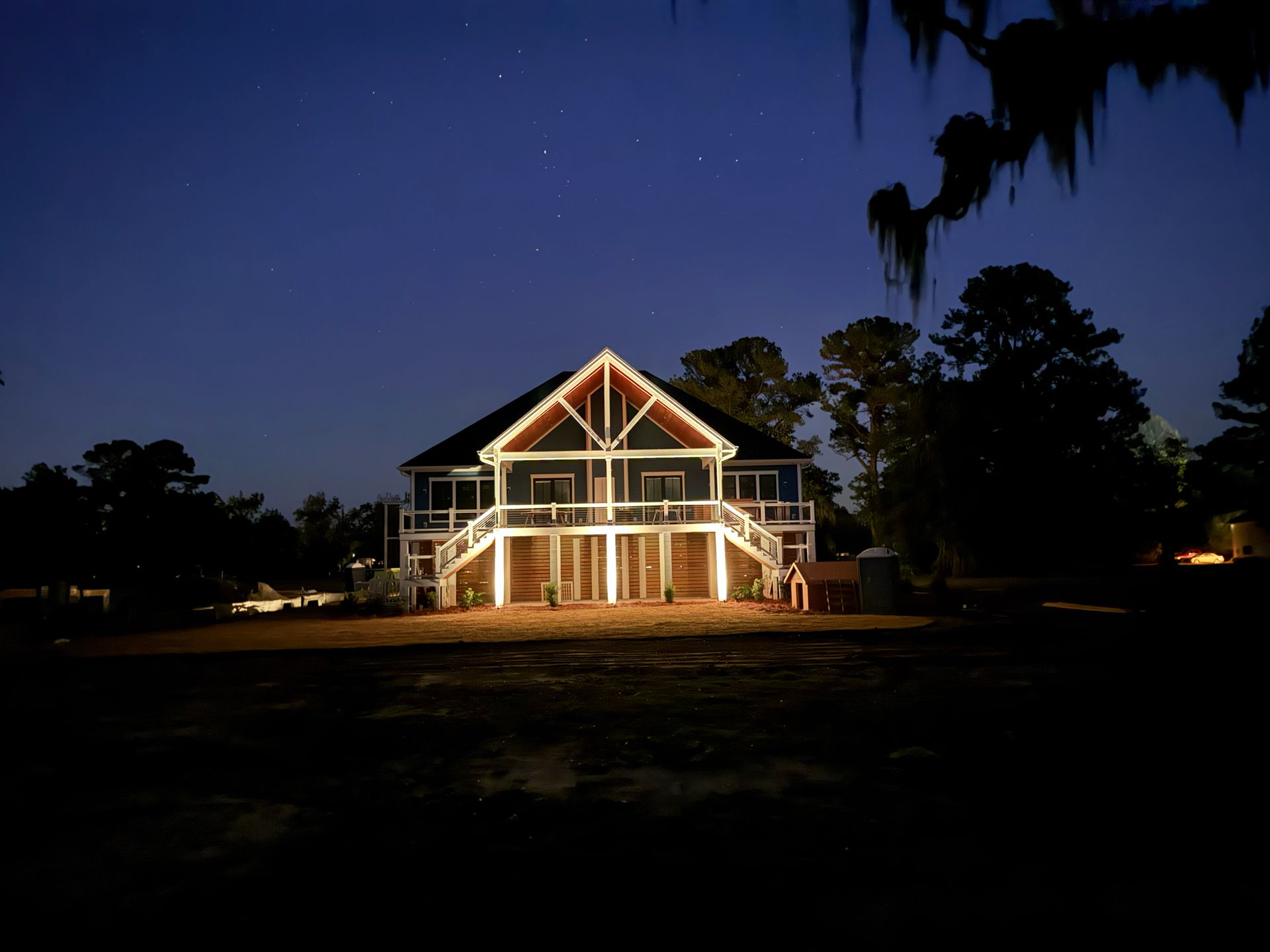 A two-story blue house illuminated at night, featuring a large wooden deck and staircase, set against a dark starry sky.