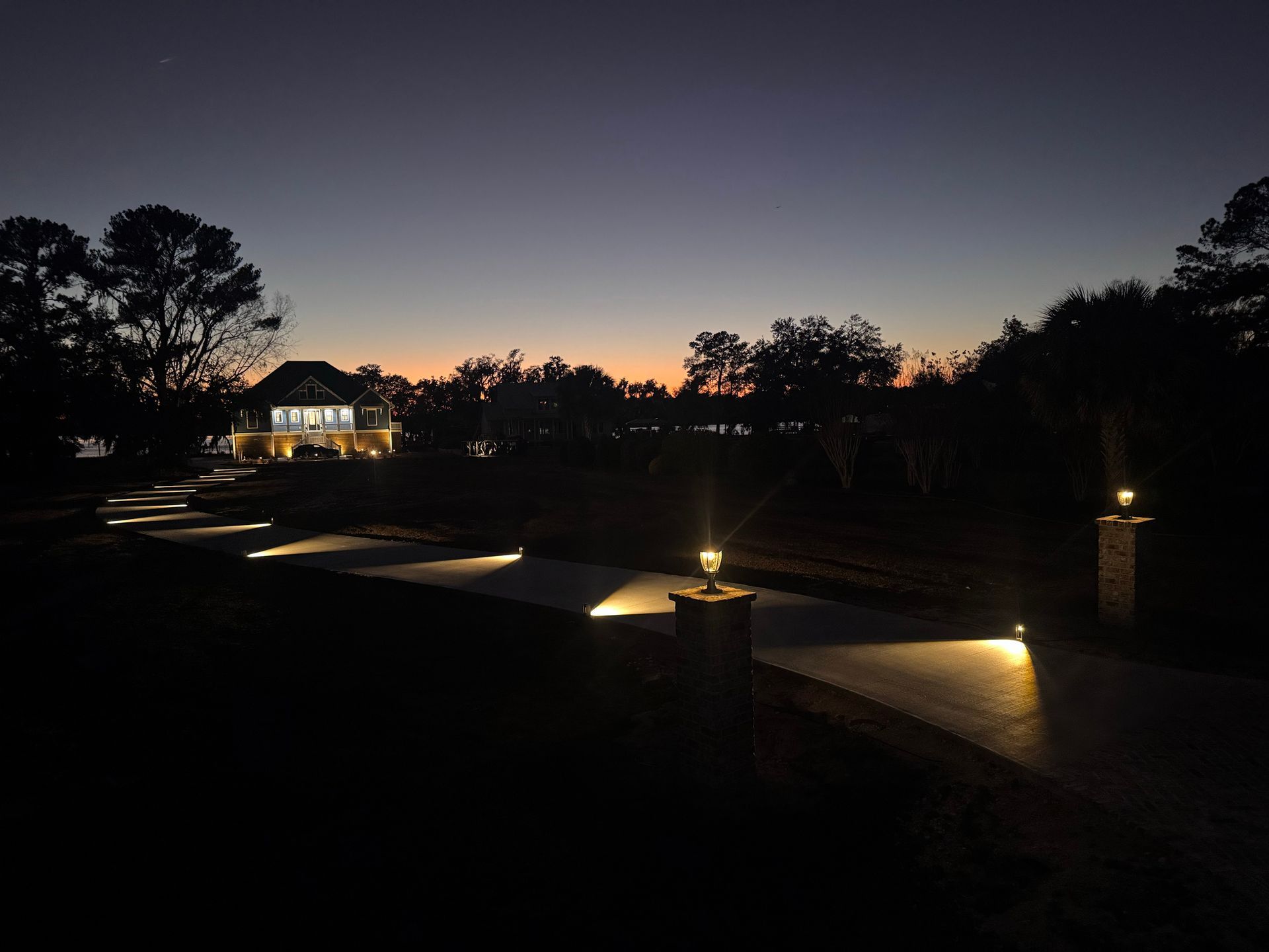 A long, winding driveway leads to a lit house at twilight, illuminated by warm post lights and glowing pathway markers.