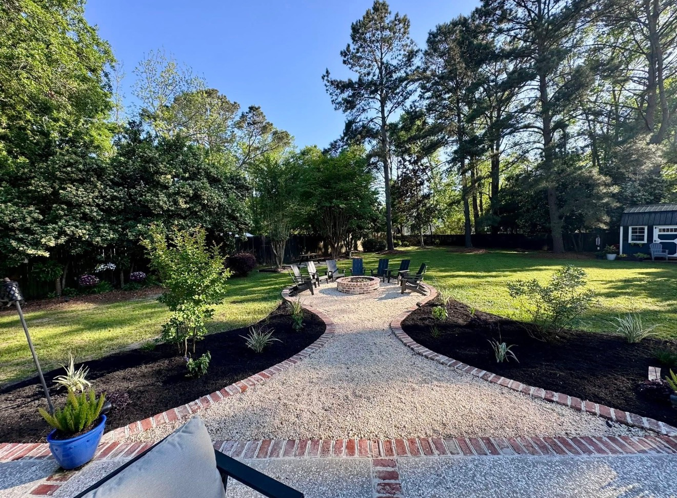 A stone-paved path with a brick border leads through a landscaped backyard to a fire pit area with chairs under trees.