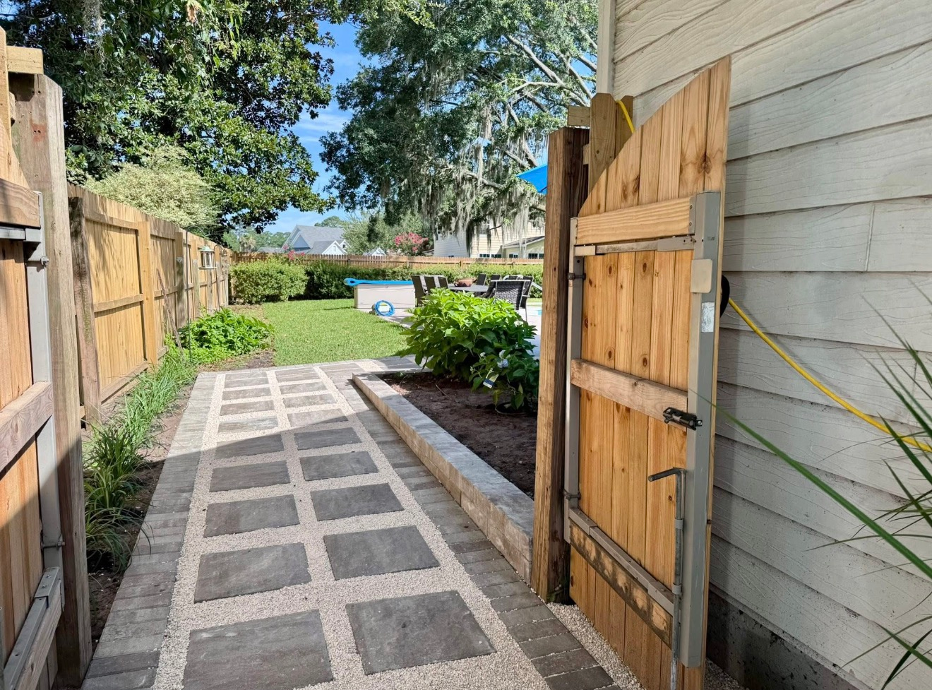 A stone-paved walkway bordered by a wooden fence and gate leads toward a backyard garden on a sunny day.