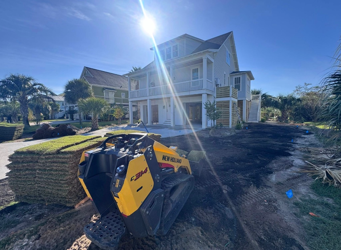 A yellow skid steer sits on a dirt lot with stacks of sod in front of a multi-story house under a bright, sunny sky.