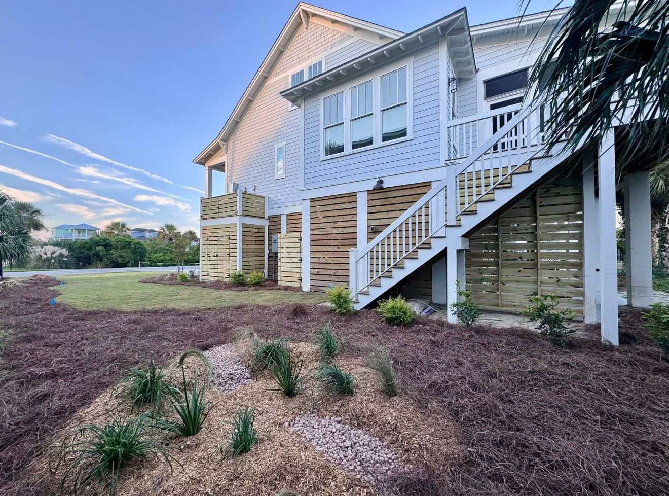 A light blue house on stilts with slatted wood paneling underneath and a wooden staircase in a mulched landscaped yard.