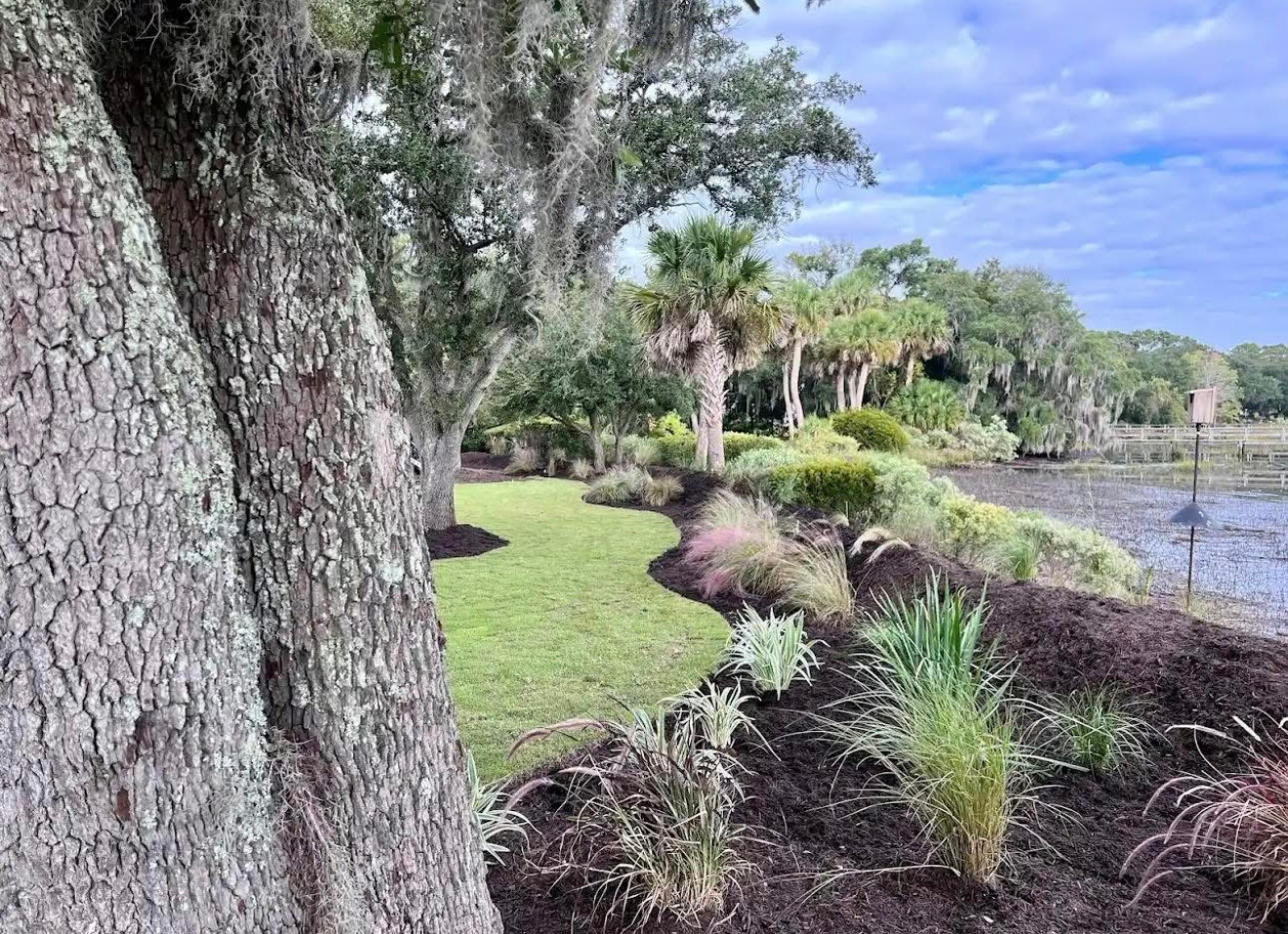A landscaped yard with a grassy lawn, mulch beds, ornamental grasses, and trees overlooking a tidal marsh.