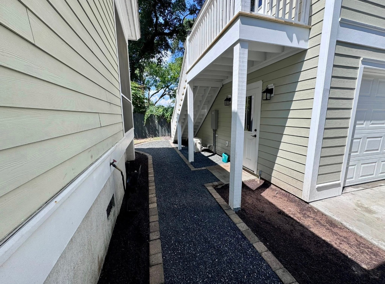 A gravel walkway between two pale yellow, wood-sided houses leads toward a sunny backyard with trees.