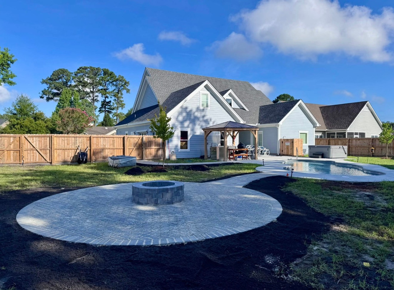 A backyard with a stone fire pit circle, a pool, and a pergola attached to the rear of a two-story residential house.