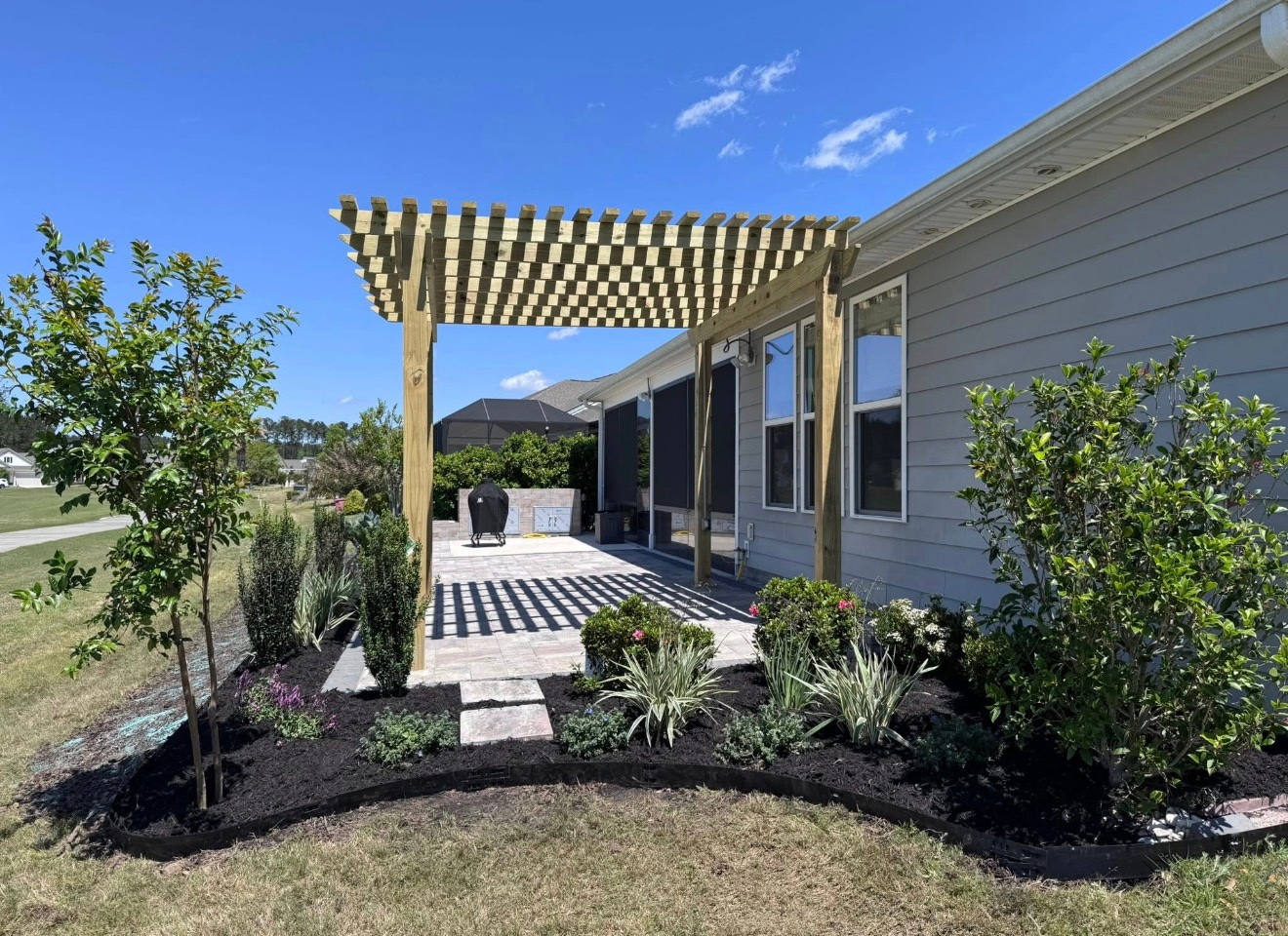 A wooden pergola covers a stone patio outside a grey-sided house, surrounded by mulched garden beds and young shrubs.