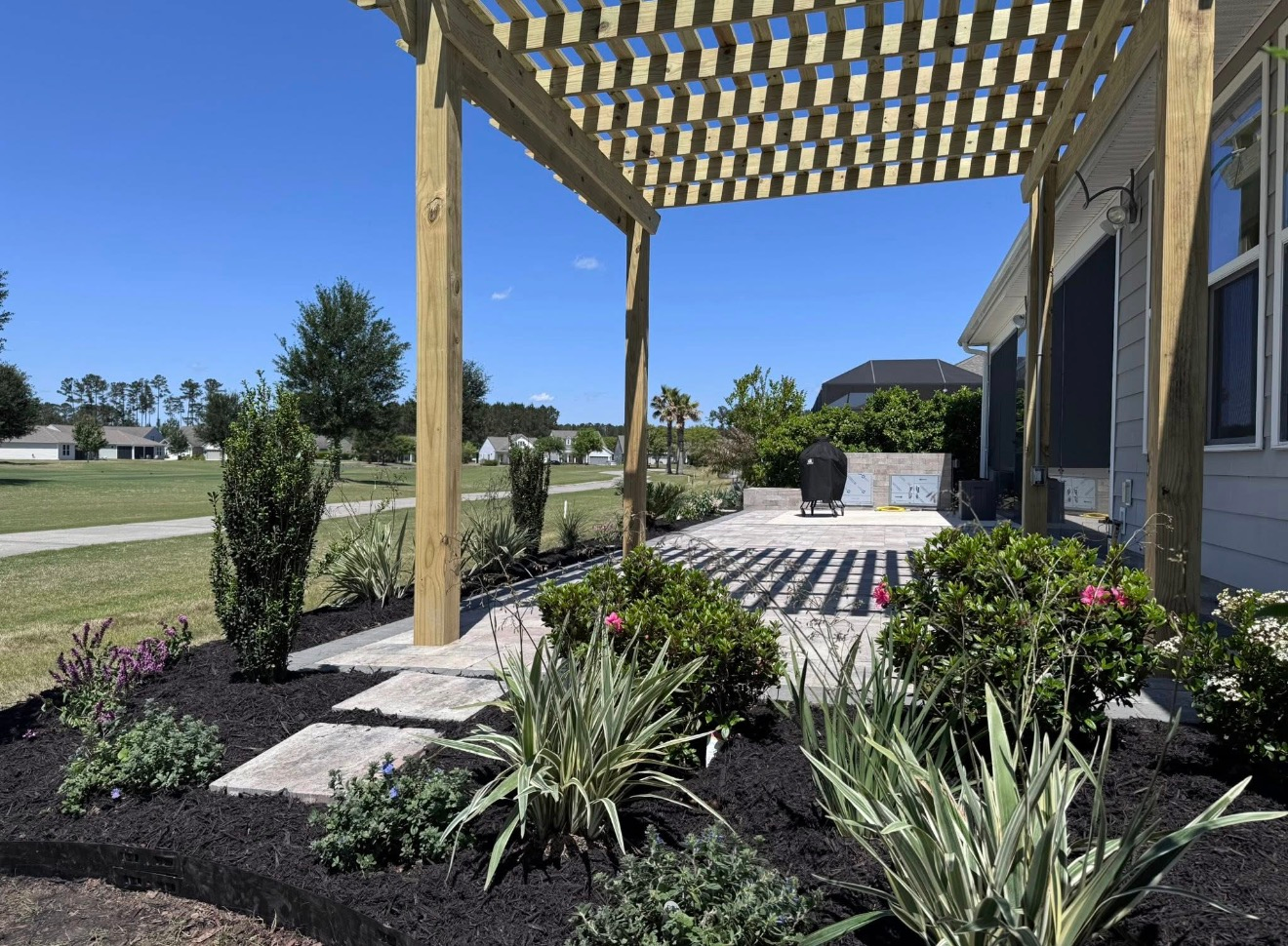 A wooden pergola frames a backyard patio with stepping stones, landscaping, and dark mulch under a bright blue sky.