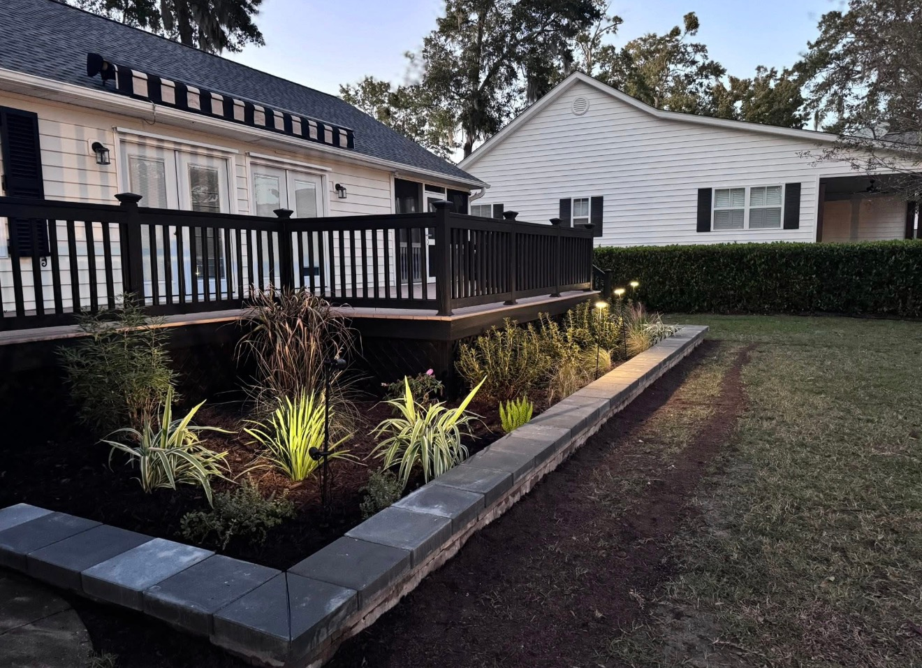 A house with a black wooden deck featuring a tiered stone garden bed planted with ornamental grasses in front.