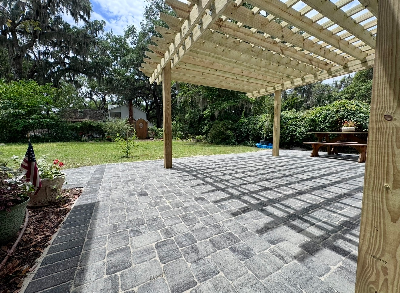 A grey paver patio with a wooden pergola, overlooking a grassy lawn with trees and a small wooden picnic table.