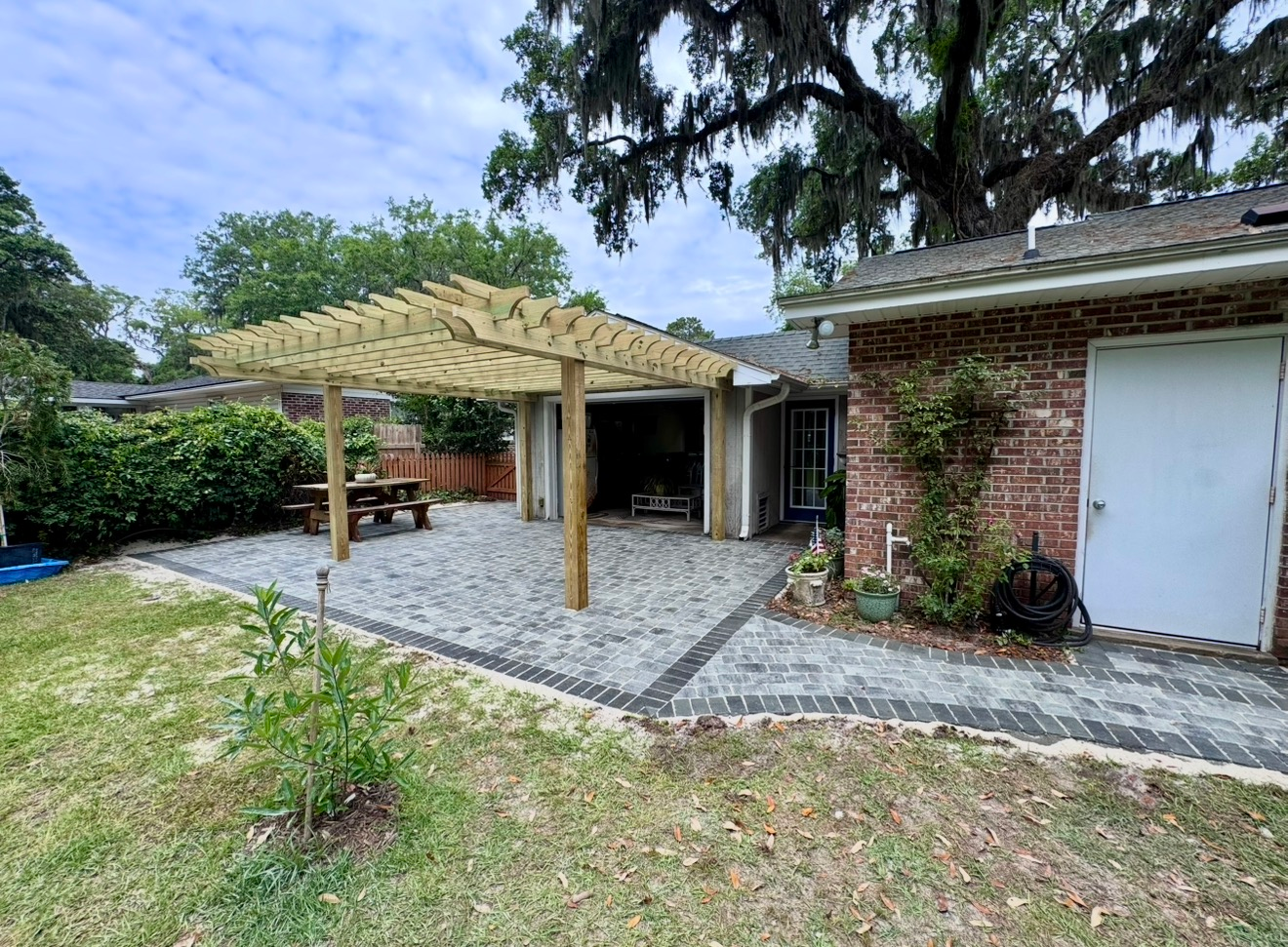 A wooden pergola stands over a gray paved patio next to a brick house with a white door and an open garage.