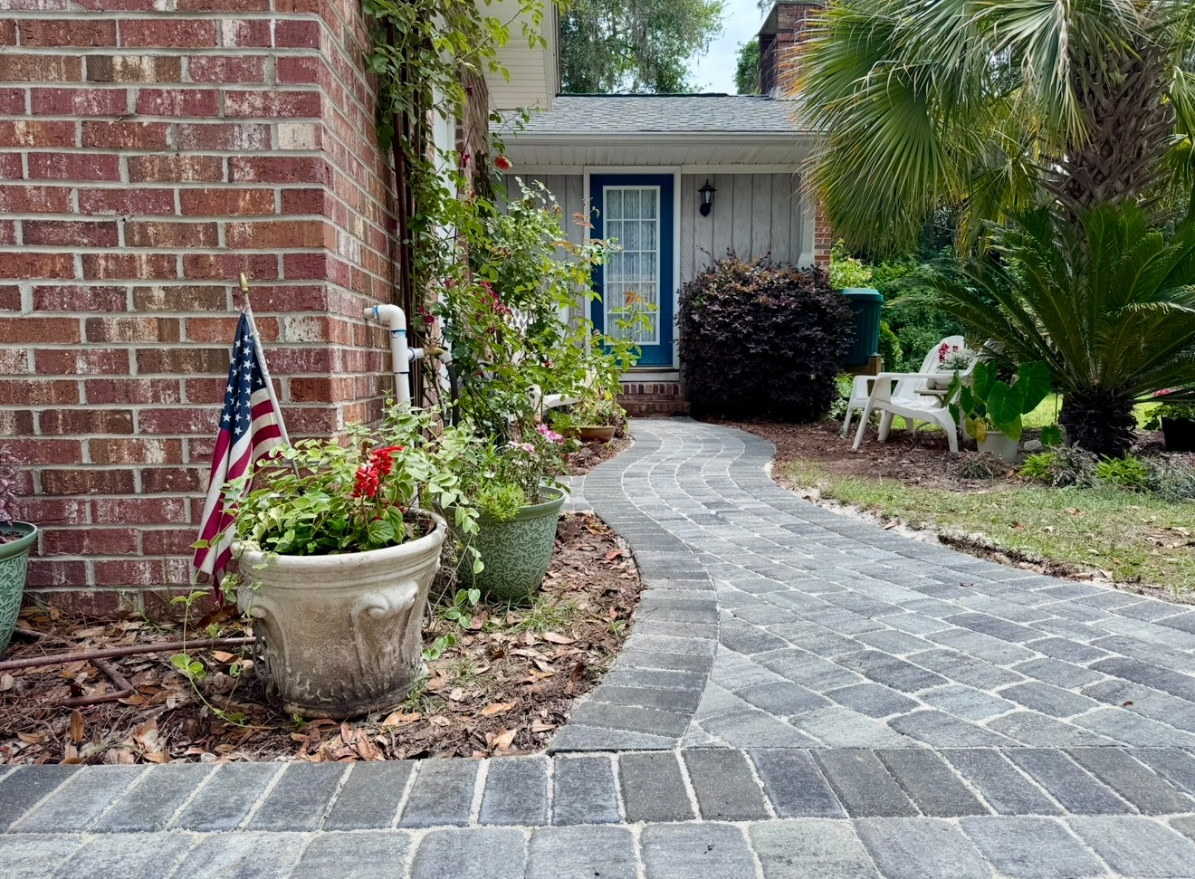 A curving gray paver walkway leads toward a blue front door, flanked by brick walls, potted plants, and lush greenery.