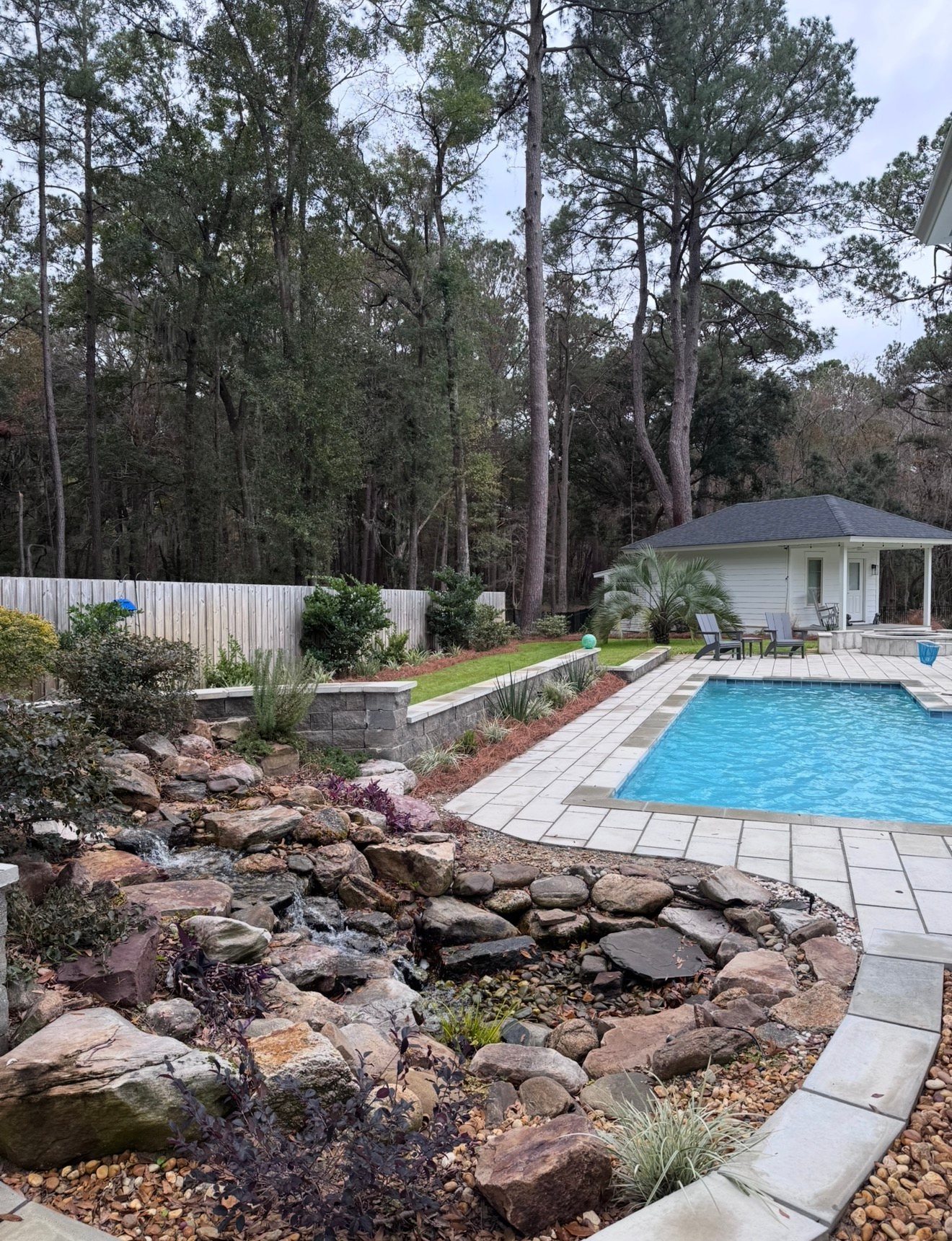 A backyard with a stone-bordered koi pond and waterfall in the foreground, next to a swimming pool and a small guest house.