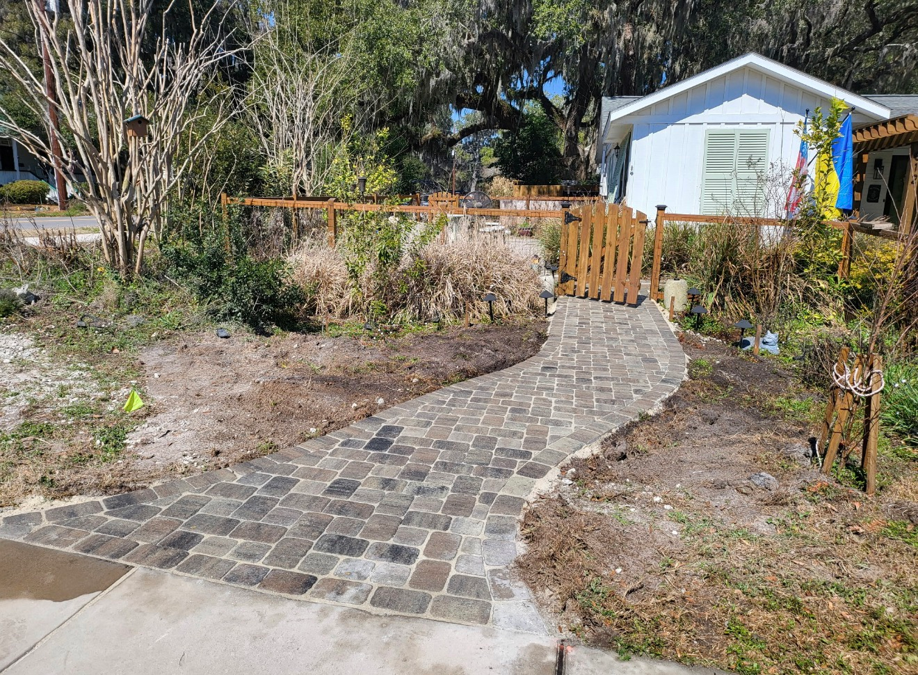 A stone path leads through a front yard toward a small wooden gate and a light-colored house in a garden setting.
