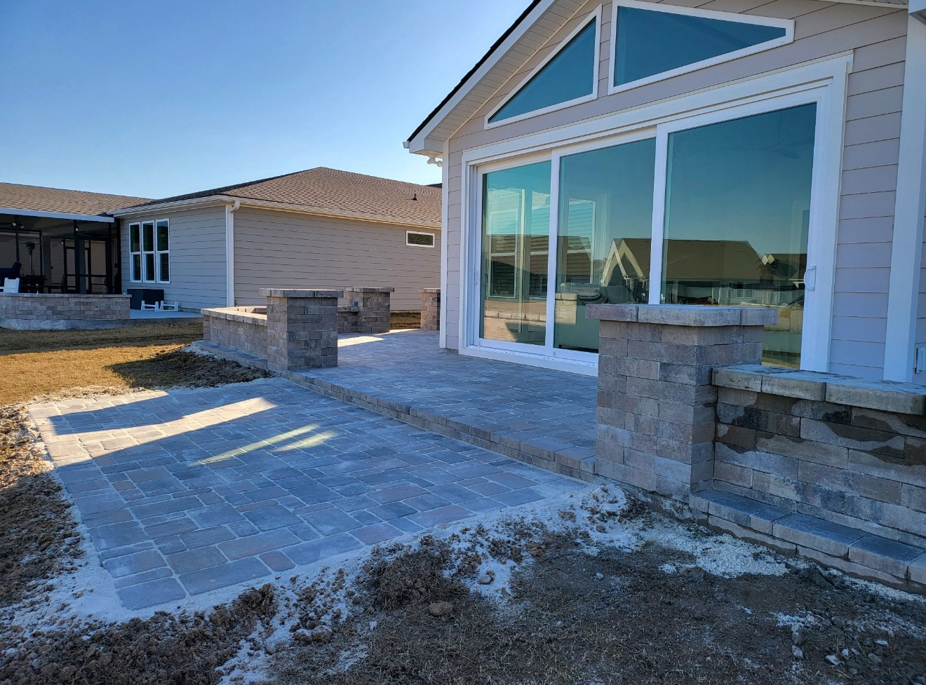 A backyard paver patio and low stone sitting wall under construction beside a light-colored house with large windows.
