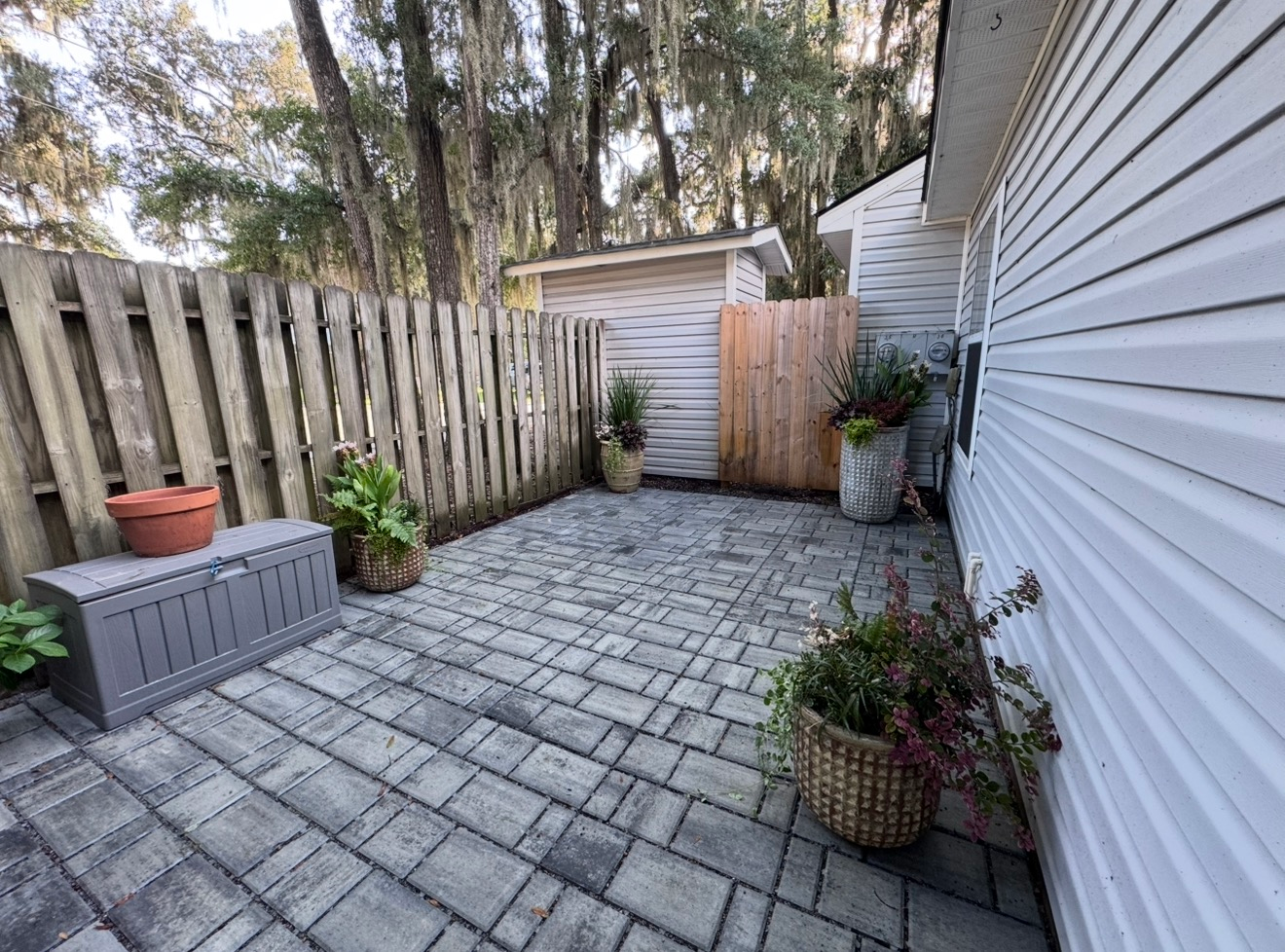 A patio with gray stone pavers, bordered by a wooden fence, a white house wall, and potted plants under leafy trees.