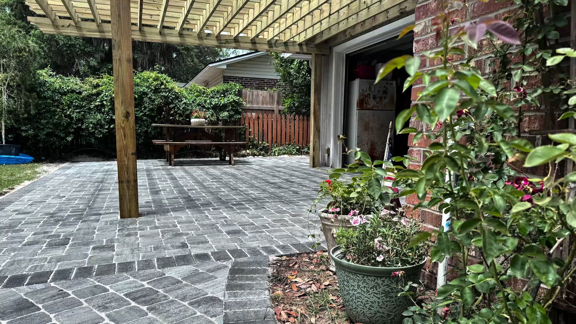 A patio with gray stone pavers, a wooden pergola, and a picnic table, viewed past a potted rose bush near a brick wall.