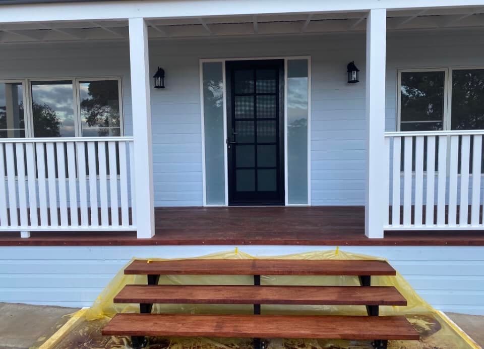 Exterior of a light blue house with a porch and steps. Dark wood steps lead up to a brown porch. Black door.