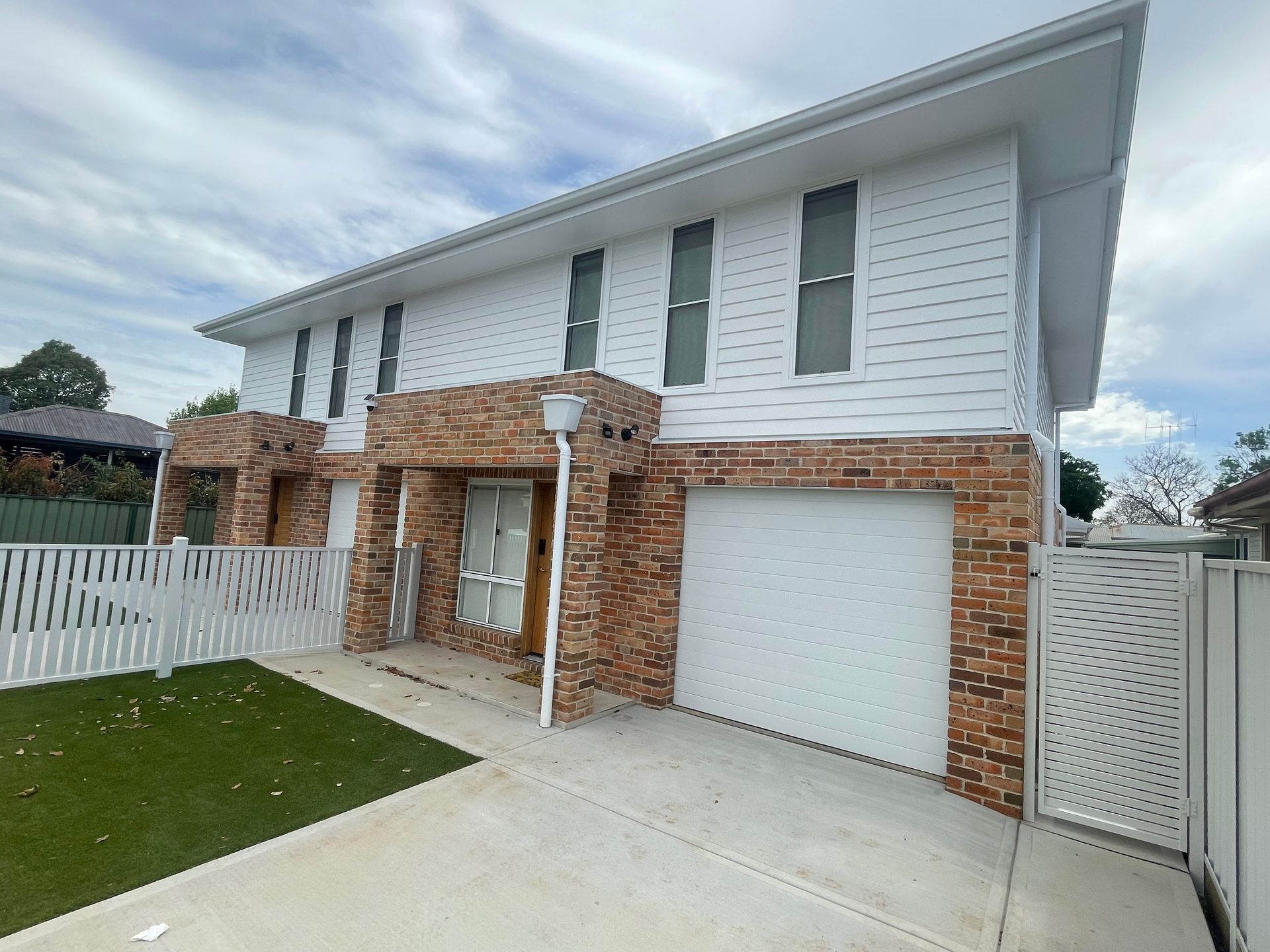 Two-story house with brick accents, white siding, garage, and decorative fence against a cloudy sky.