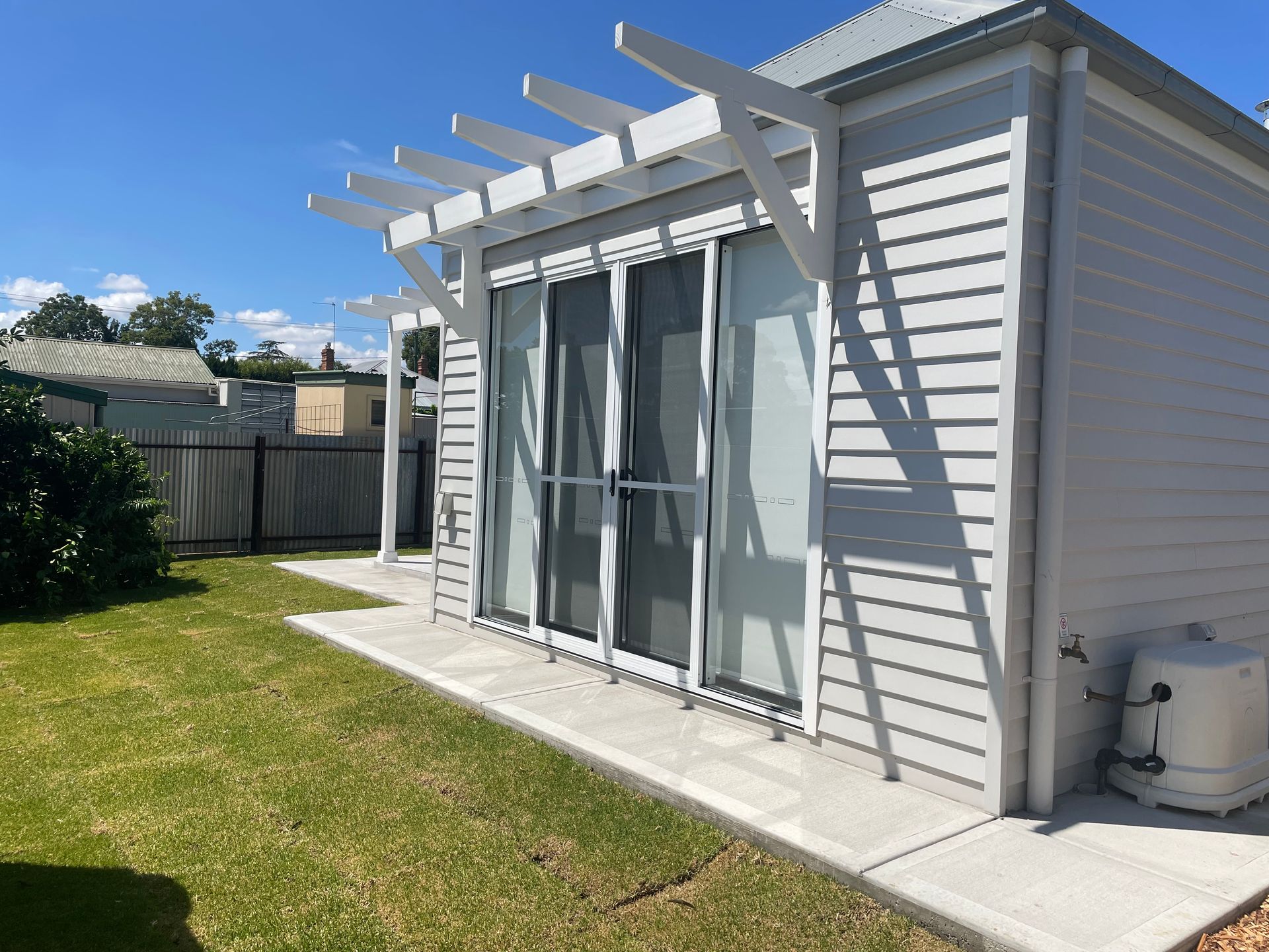 Light gray building with sliding glass doors, white pergola, concrete patio, and green grass.