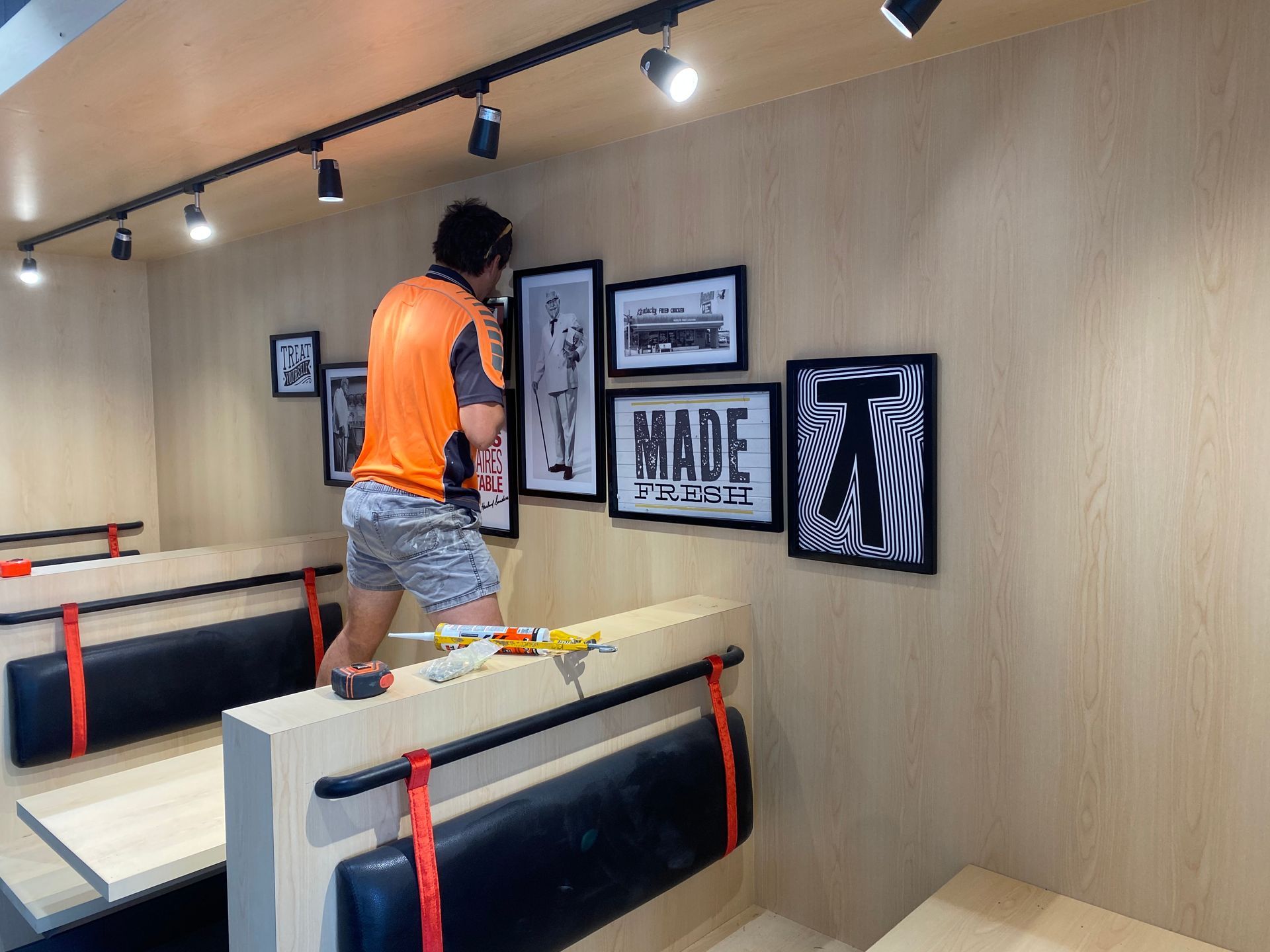 Man hanging framed artwork on a wood panelled wall above booth seating, lit by track lighting.