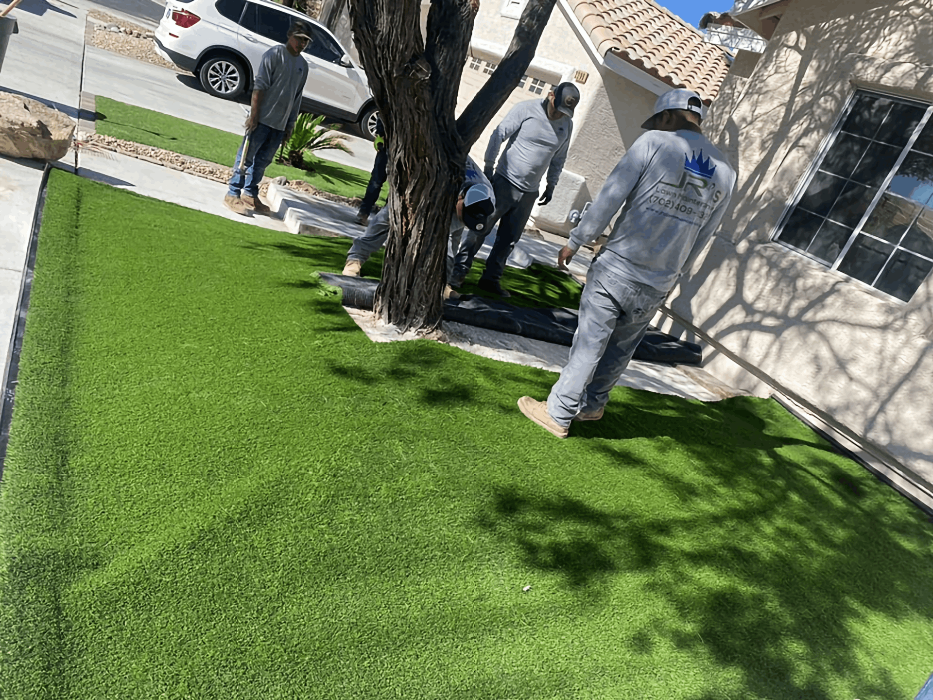 Workers installing artificial grass in a front yard beside a house and tree.