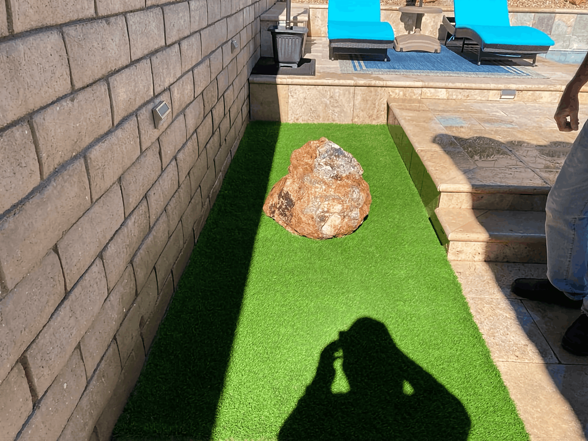 Outdoor patio with artificial grass, a large rock, lounge chairs, and a wall shadow in sunlight