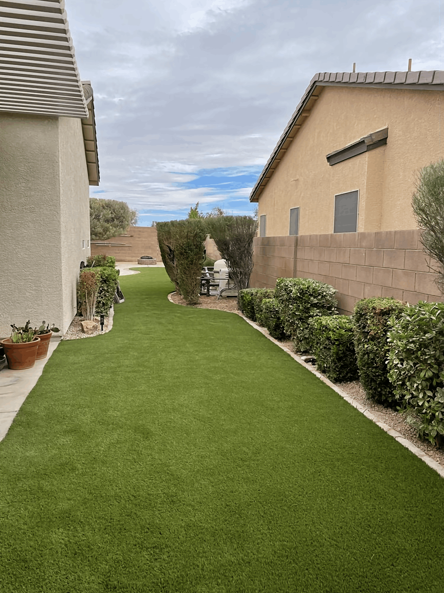Narrow green lawn path between two houses, bordered by hedges and potted plants under a cloudy sky