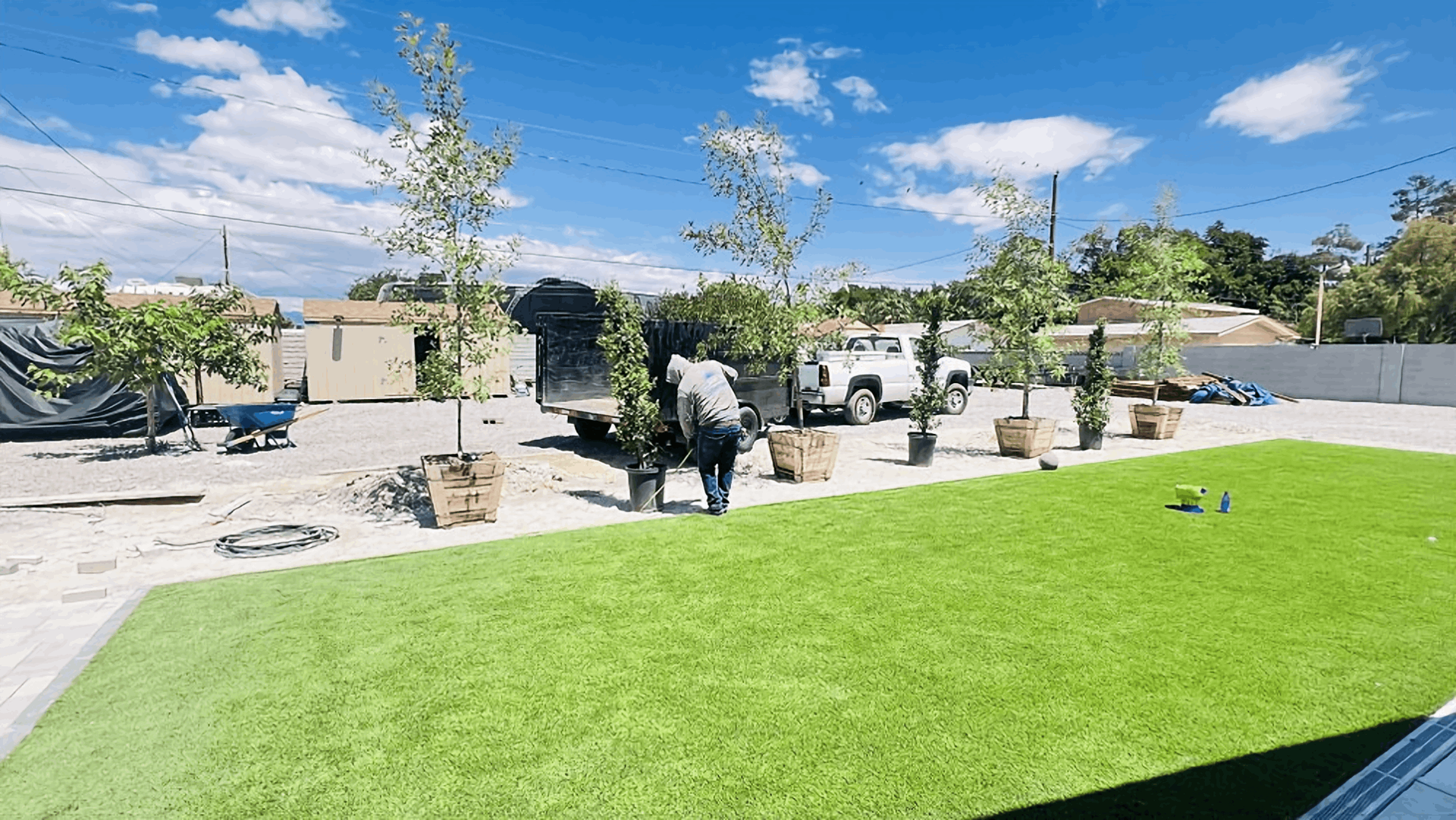 Sunny backyard with bright green lawn, patio seating, potted trees, and a blue sky with scattered clouds