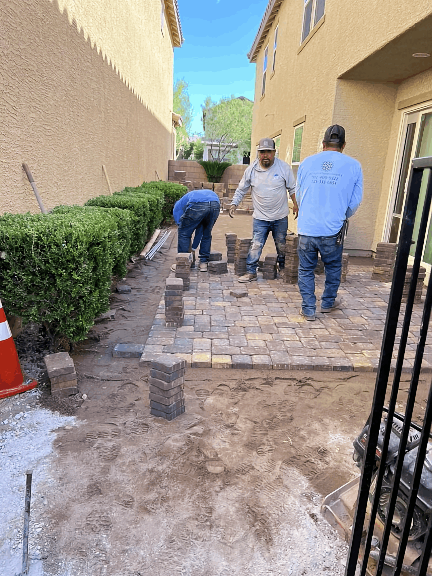 Workers laying a brick walkway beside a beige building, with pavers, dirt, and a hedge along the path.