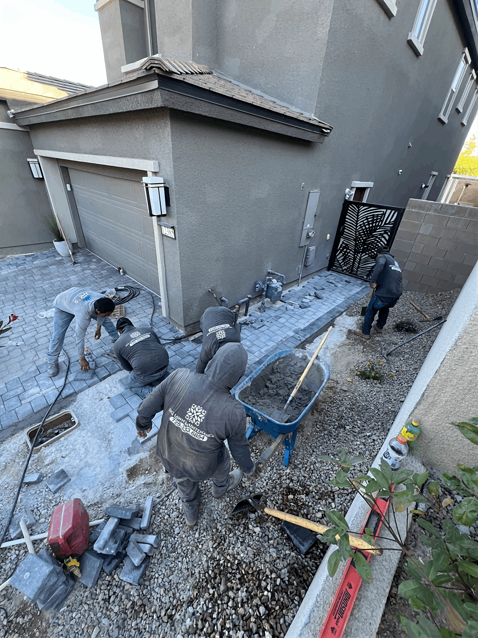 Workers pouring a concrete walkway beside a house, with wheelbarrows and tools in a narrow yard