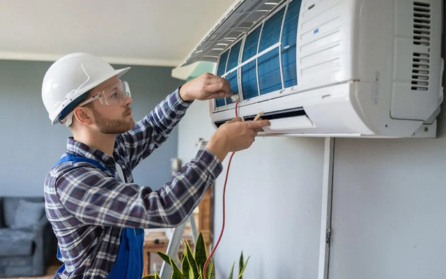 HVAC technician in a hard hat and safety glasses, working on a wall-mounted AC unit.
