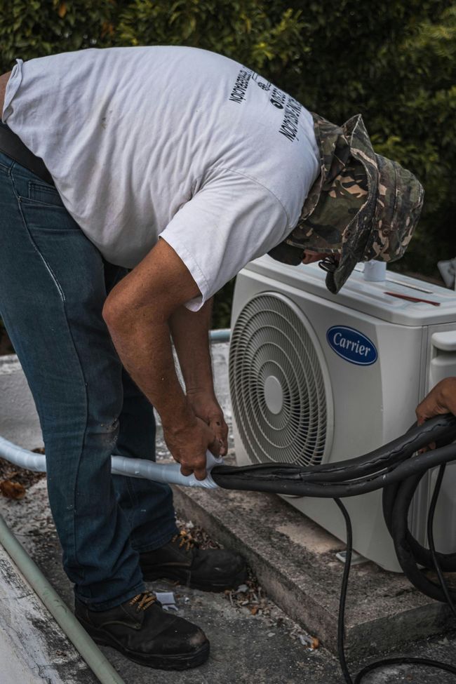 Person working on an air conditioning unit outside. He wears a white shirt, jeans, and a camouflage hat.