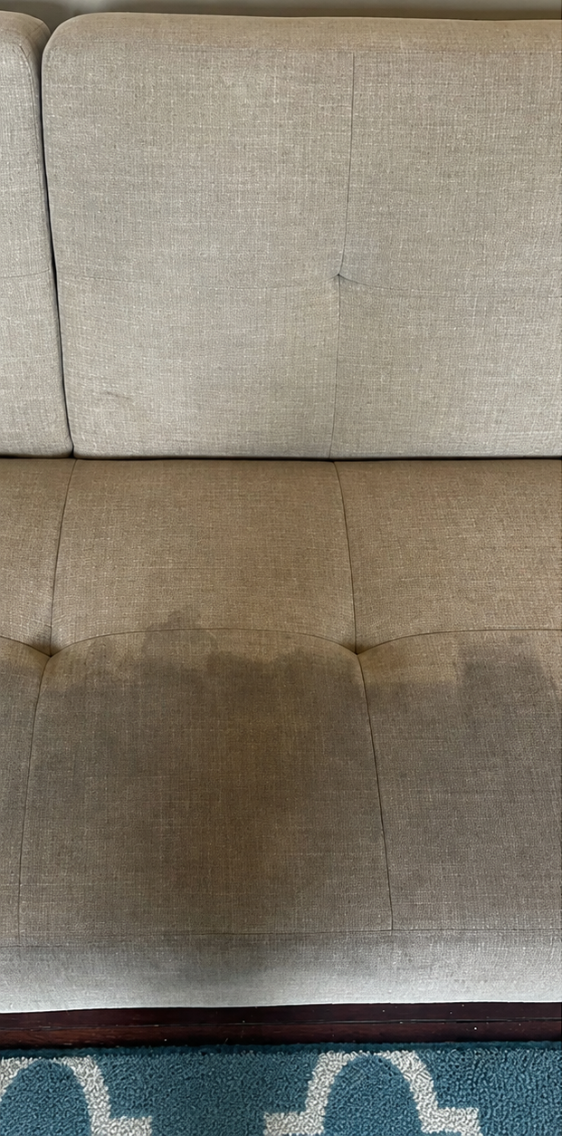 Close-up of a beige upholstered sofa cushion with a dark stain in the center