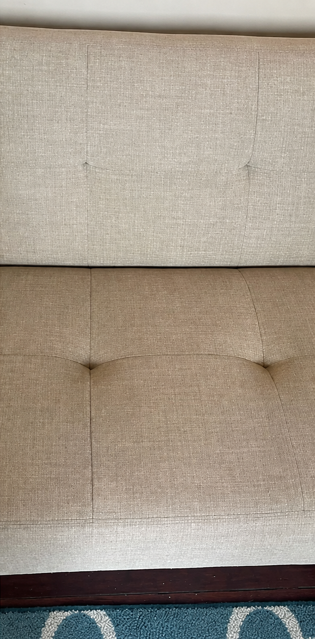 Close-up of a beige upholstered sofa with tufted back cushions and a dark wooden trim at the bottom