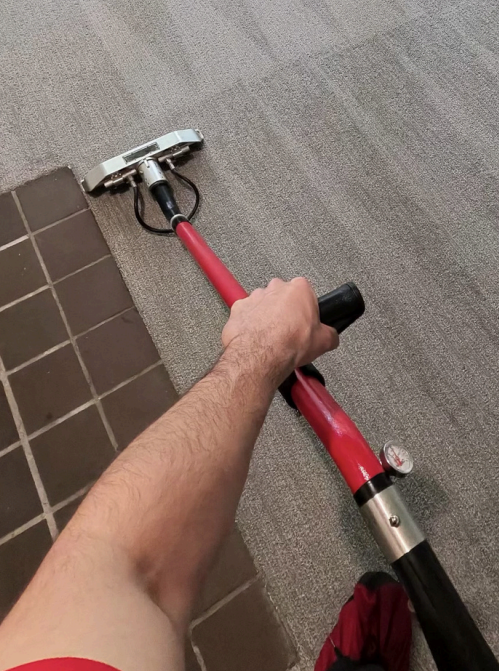 Person using a red-handled floor scrubber on gray carpet next to brown tile flooring