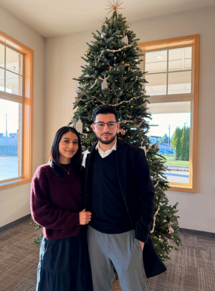 Two people standing indoors in front of a decorated Christmas tree.