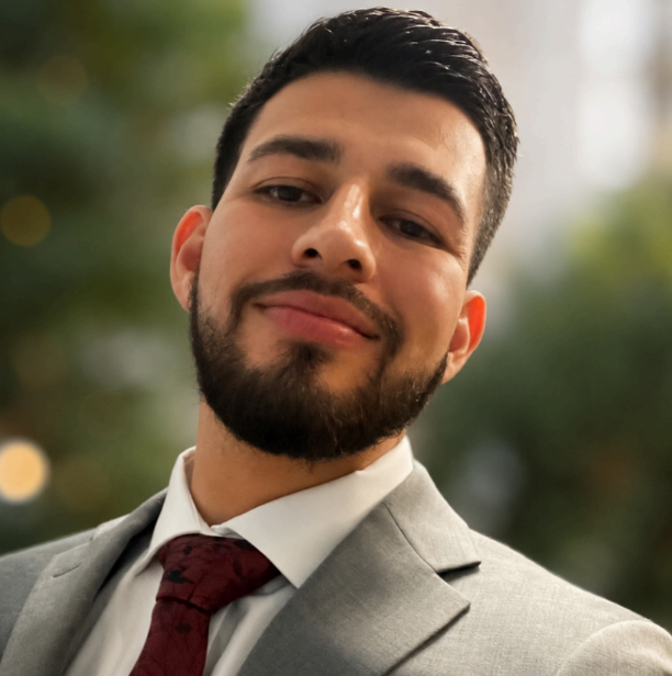 Man in a gray suit and burgundy tie, smiling against a blurred outdoor background
