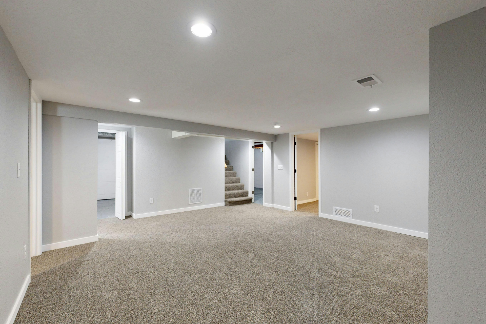 Empty carpeted basement room with gray walls, recessed lights, and a staircase in the center background