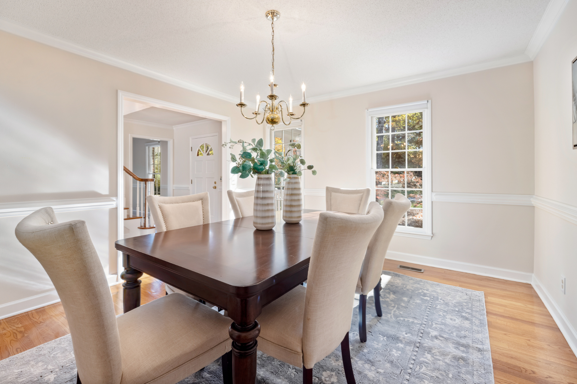 Bright dining room with wooden table, cream chairs, chandelier, and large window on hardwood floors